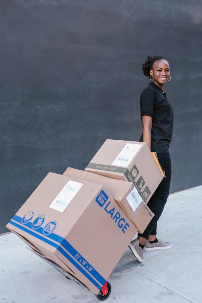 Young woman delivering cardboard boxes on a dolly outdoors, smiling confidently.