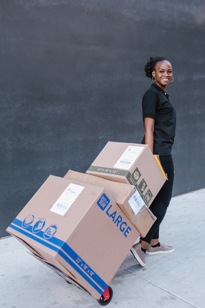 Young woman delivering cardboard boxes on a dolly outdoors, smiling confidently.